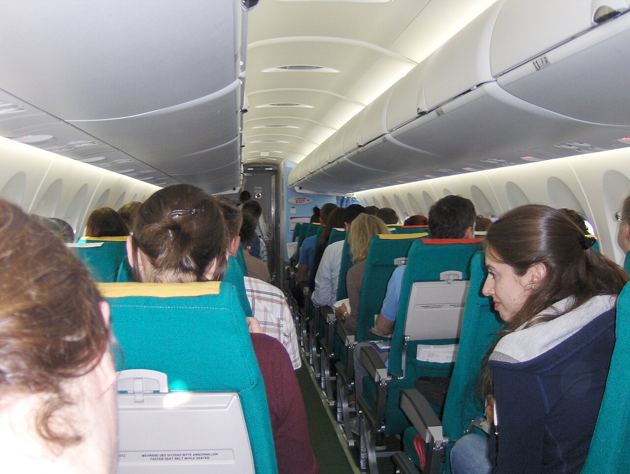 The inside of an airplane cabin, looking down the aisle past rows of seats, with the overhead bins above