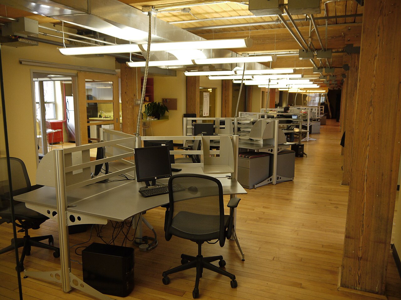 A bright open-plan office with rows of desks, computers, and low cubicle dividers