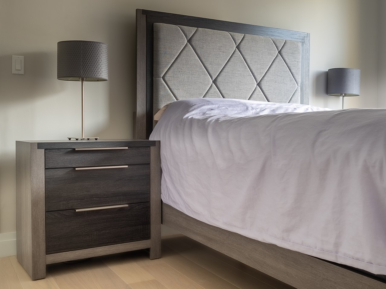 A made bed with a padded headboard and comforter, next to a nightstand with a lamp and drawers