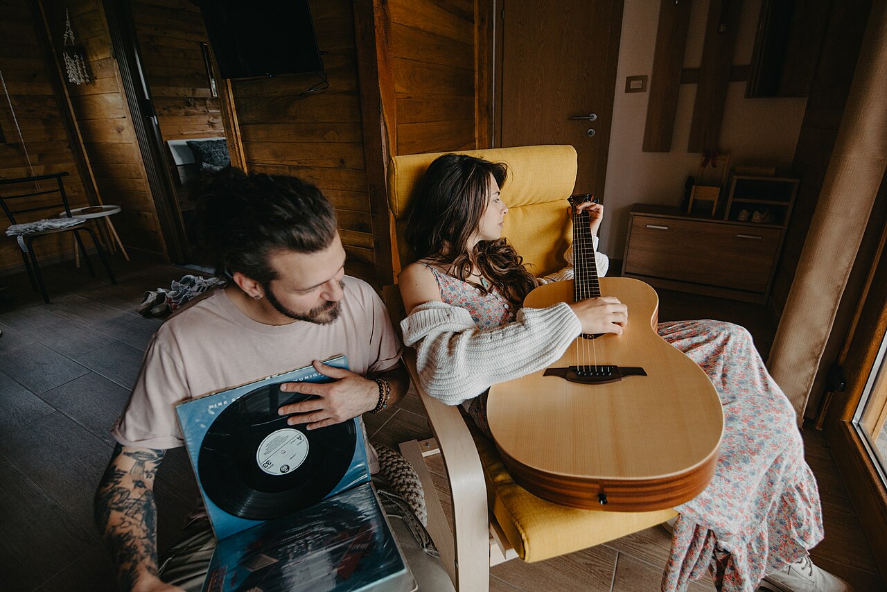 A young couple relaxing together in a cabin, one playing an acoustic guitar and the other holding a vinyl record