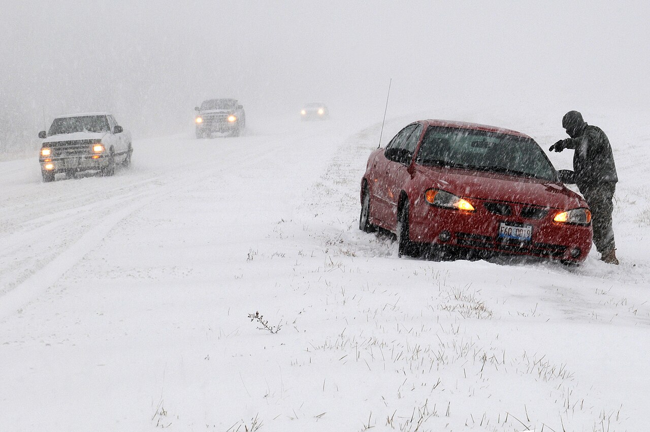 A red car stuck at the roadside during a heavy blizzard, with snow blowing across the highway